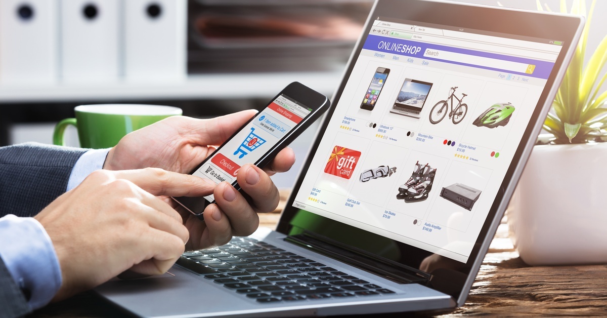 A man's hands hold his smartphone in front of his laptop. Both screens are showing online shopping pages.
