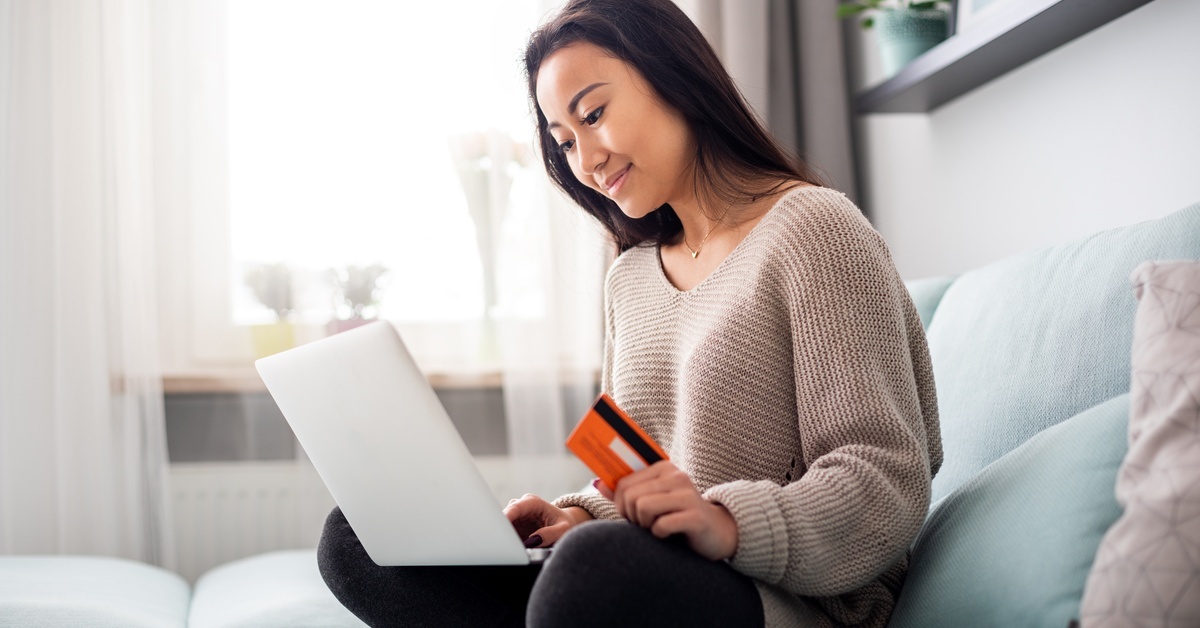 A woman holding a credit card in one hand sits on her couch in a sweater looking at the screen of her laptop.