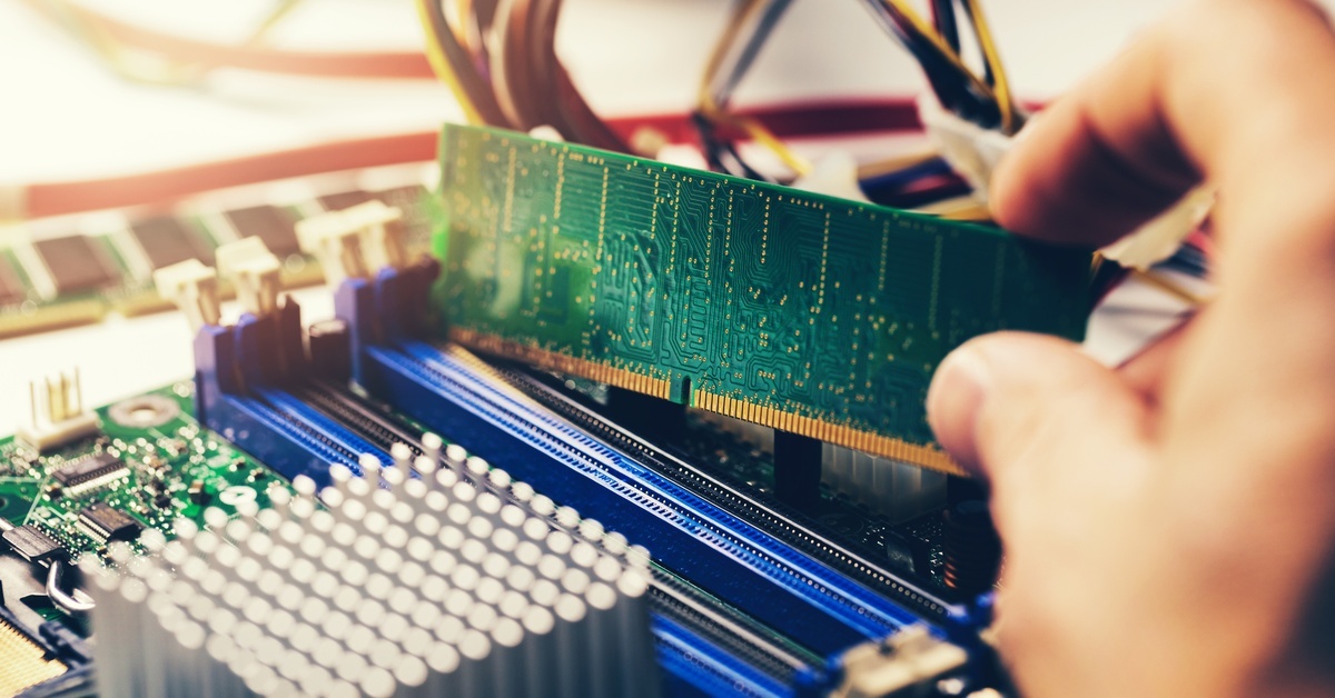 An engineer's hand is placing a green random access memory module into the motherboard of an open desktop computer.