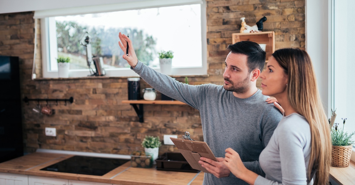 A couple standing in the kitchen of their home. The man holds a clipboard as he gestures to the rest of the room.