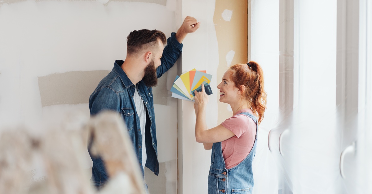 A couple standing in a room under construction. The woman holds up multiple paint swatches to the man next to her.