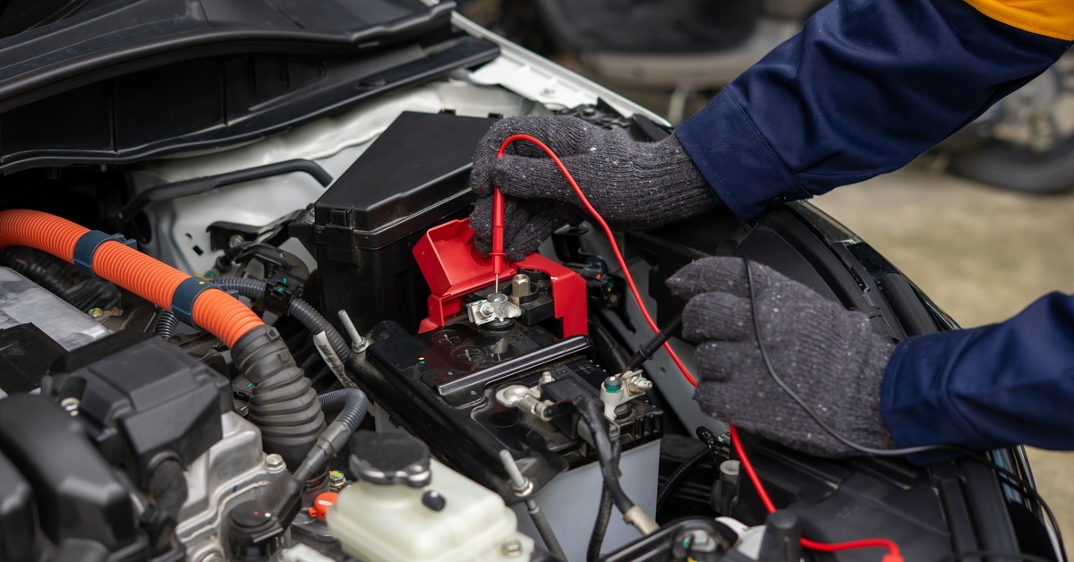 A mechanic wearing gloves uses a multimeter to test the charge of a car battery in the open hood of the car.