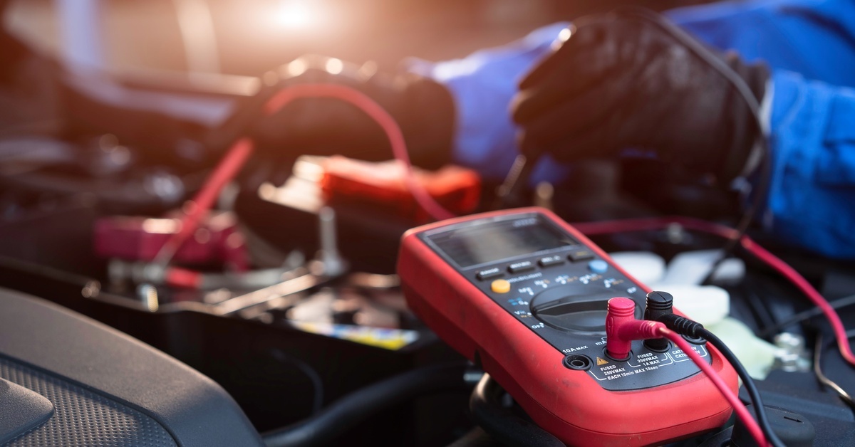 A multimeter with red and black wires coming out of it is being used by a technician on a car battery.