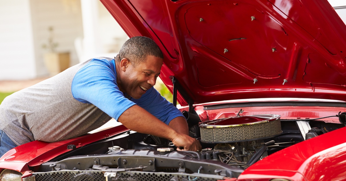 A man working on his car with the hood open. He is smiling as he reaches into the engine of the vehicle.