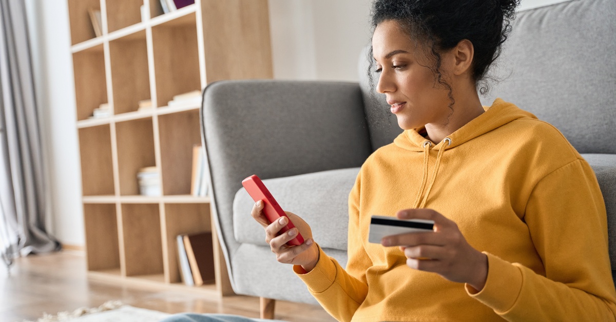 A young woman sitting on the floor and looking at the phone in her hand. Her other hand is holding her credit card.