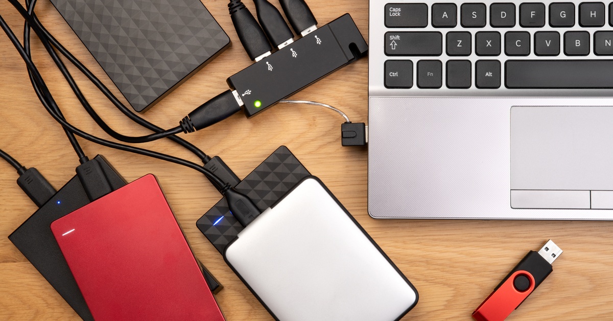 A series of external hard drives connected to a silver laptop. A small thumb drive is in front of the laptop.