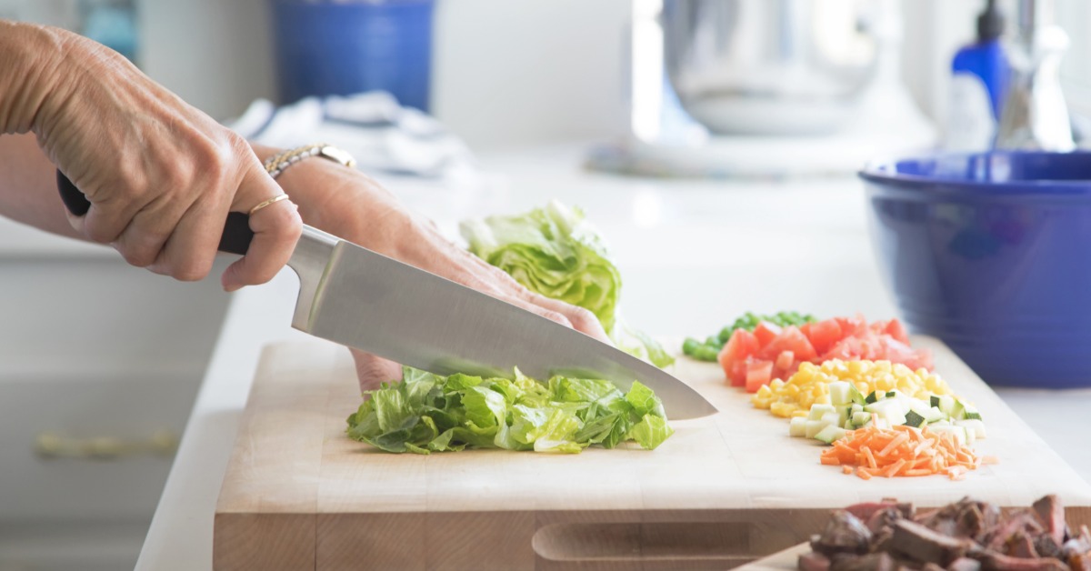 A woman's hands using a knife to chop up lettuce on a cutting board. The board already has several other prepared vegetables.