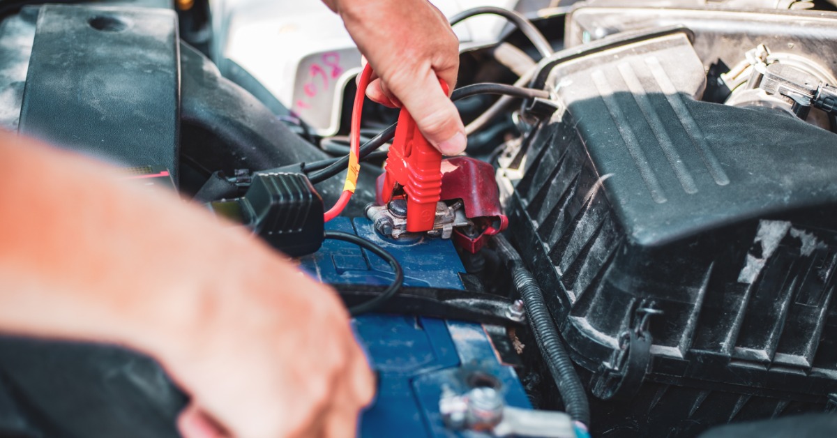 Beneath an open car hood, a pair of hands uses red and black spark plugs to connect to the car's battery ports.