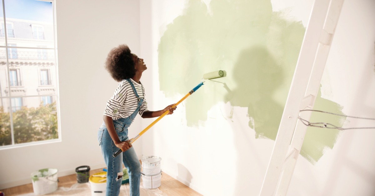 A woman wearing overalls in a half-finished room using a paint roller to roll a layer of green paint onto a white wall.