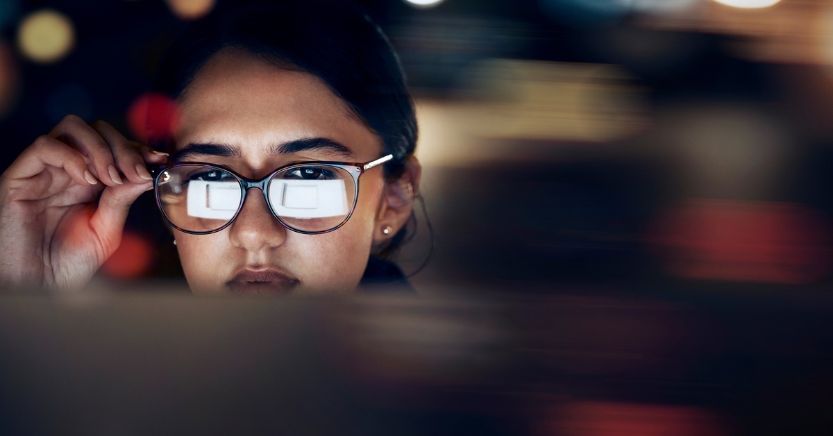 A woman holding her hand up to the side of her glasses. A computer screen is reflected in her glasses.