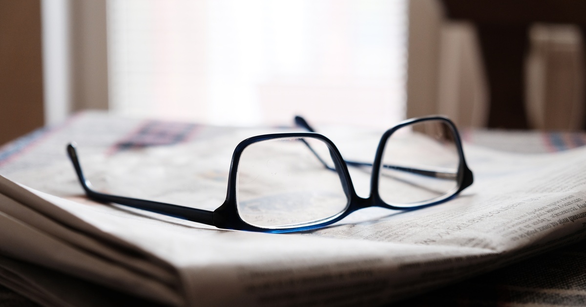 A pair of glasses laying upside down on a folded up newspaper. There is a sunlit window behind the glasses.