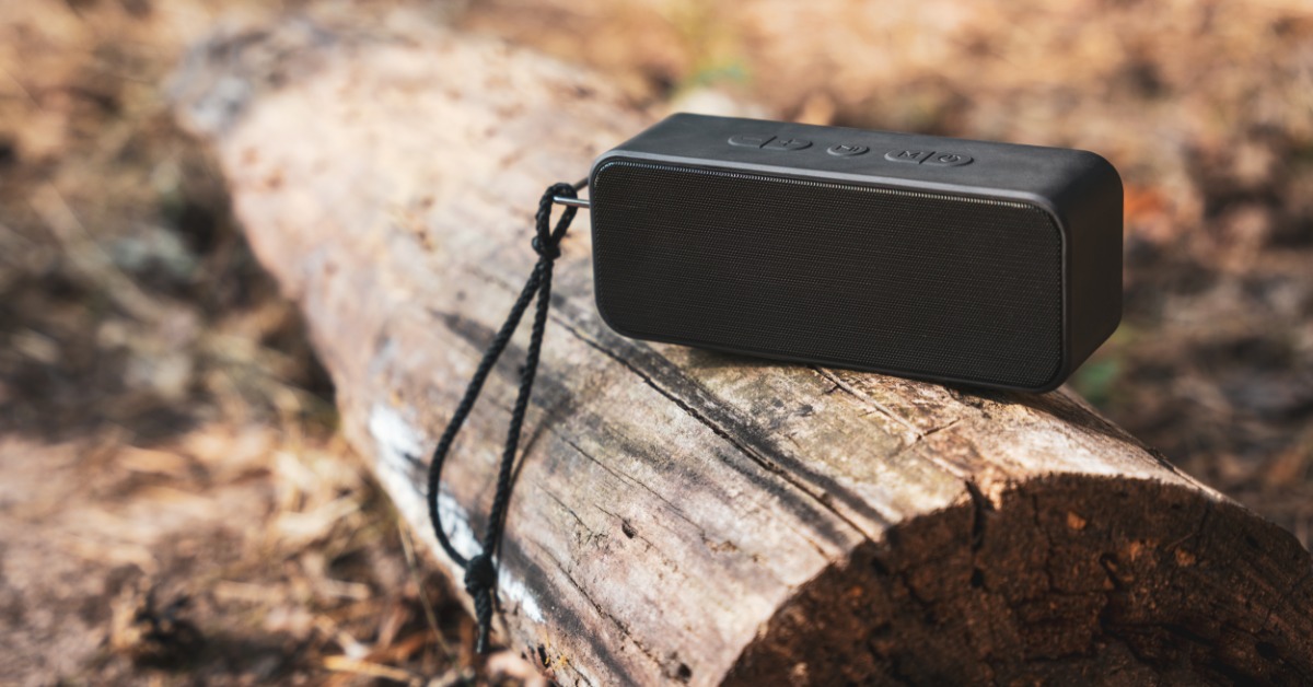 A portable, wireless, Bluetooth speaker sitting outside on a log that is partially embedded in the ground.