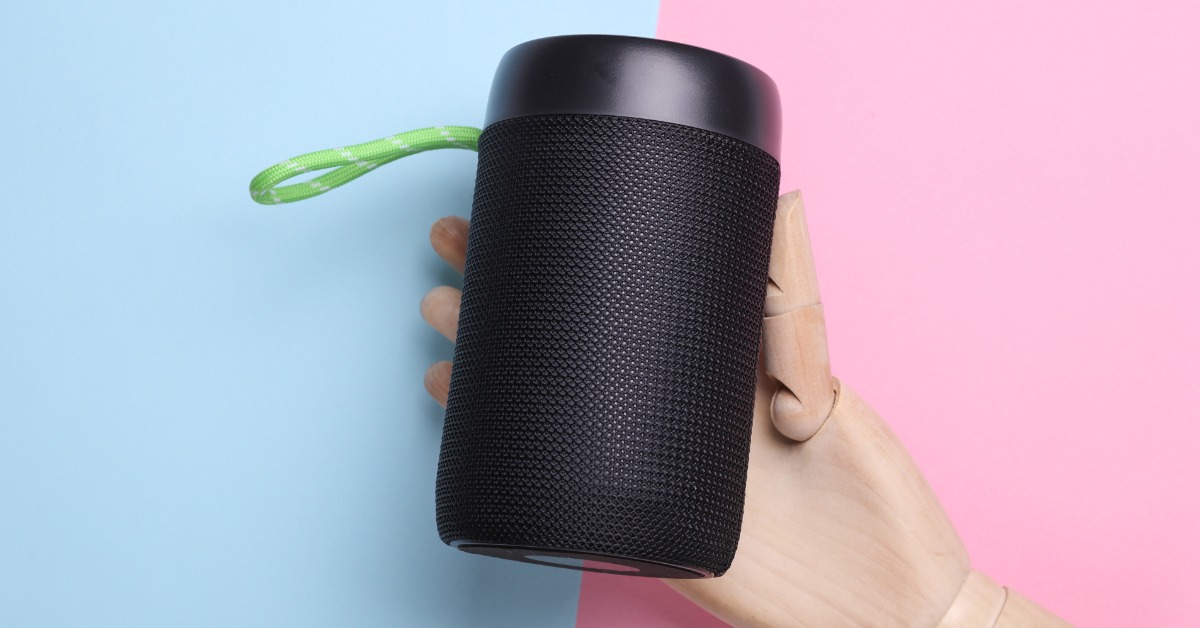 A wooden puppet hand holding a Bluetooth speaker. The hand is up against a half pink and half blue background.