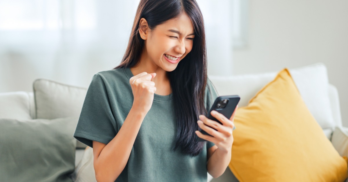 A woman sitting on her couch with her phone and credit card in hand. She is smiling down at her phone.
