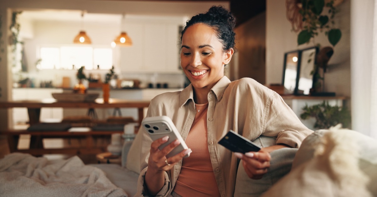 An older woman sitting on a couch in front of her laptop. She is holding her credit card and cheering.
