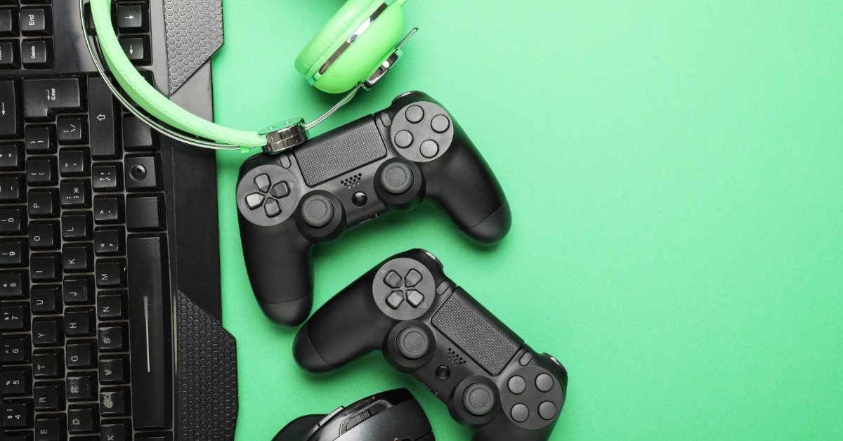 Two gaming controllers, a mouse, a keyboard, and a pair of headphones all laying in a pile against a green background.
