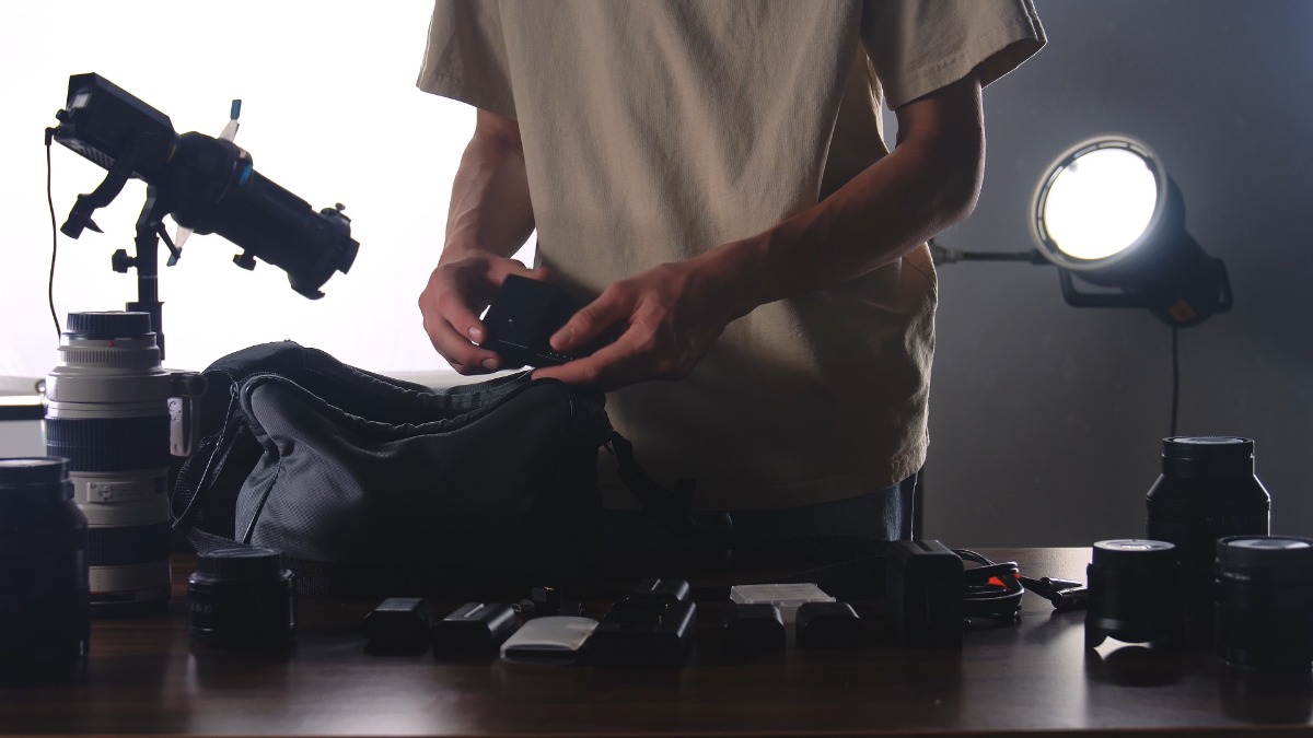How To Avoid Overpaying for Digital Camera Accessories A man organizing a bunch of camera equipment on a table. There is a spotlight illuminating him from behind.
