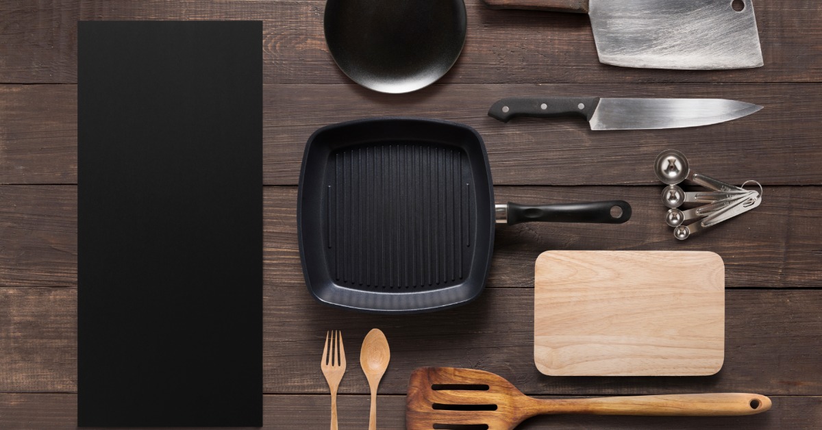 A neatly arranged variety of kitchen tools on a wooden table. A large pan and chef's knives are placed next to each other.