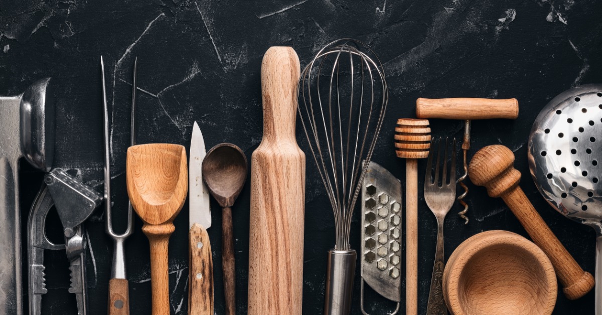 A lineup of various kitchen utensils on a marble counter. Knives, whisks, spoons, and other utensils make up the line.